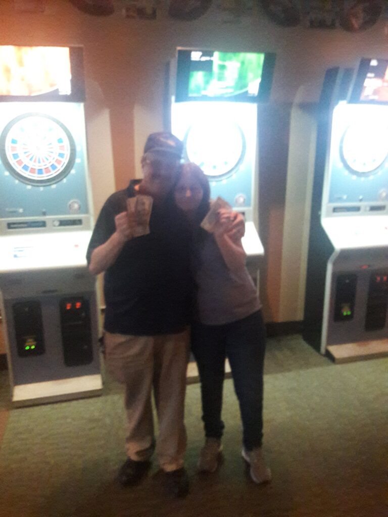 Man and woman hugging and holding up cash in front of the dart boards where they won a tournament