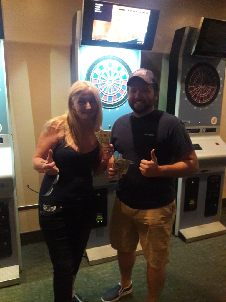 A lady and a man in front of a dart board giving the thumbs up after winning cash