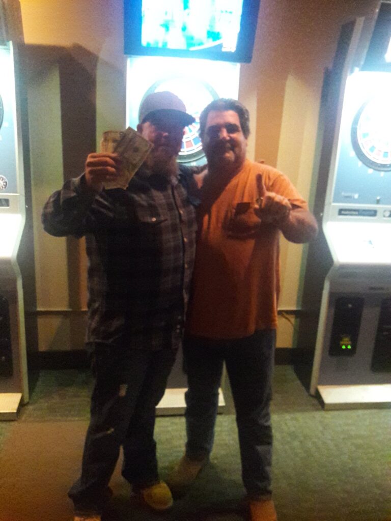 Man in flannel shirt holding up cash next to a man in a tshirt holding up one finger after winning at darts